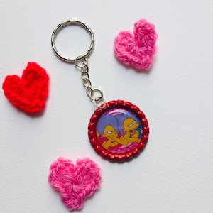 May include: A red bottle cap keychain featuring a picture of two cartoon characters. The keychain is surrounded by three small crocheted hearts, two pink and one red.
