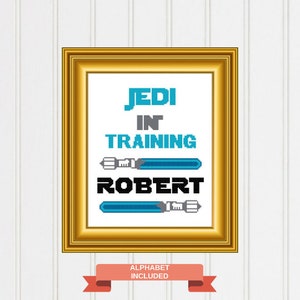 May include: A framed cross-stitch design with the text "Jedi in Training Robert" and two blue lightsabers. The frame is gold and the background is white.