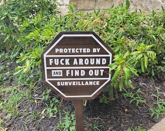May include: A brown wooden sign with white text that reads "Protected by Fuck Around and Find Out Surveillance". The sign is shaped like an octagon and is mounted on a wooden post.
