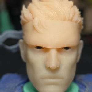 Op de afbeelding: Een close-up van een lichtgekleurde actiefiguurkop met gedetailleerde gelaatstrekken, waaronder een gefronste wenkbrauw en een strenge uitdrukking. De figuur heeft kort, stekelig haar en een hoofdband. De figuur staat tegen een vage achtergrond.