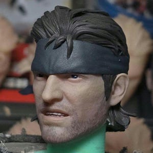 Könnte beinhalten: Detaillierte Kopfskulptur eines männlichen Charakters mit kurzen, dunklen Haaren und einem schwarzen Stirnband. Die Figur hat einen Bartstoppel und einen entschlossenen Gesichtsausdruck. Der Hautton ist realistisch.