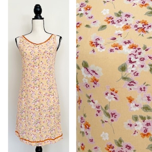 Könnte beinhalten: Ein ärmelloses, gelbes Blumenkleid mit V-Ausschnitt und orangefarbenem Besatz. Das Kleid hat ein kleines Blumenmuster mit rosa, orangefarbenen und weißen Blumen.