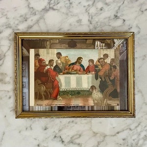 Op de afbeelding: Een ingelijste vintage print van een schilderij dat het Laatste Avondmaal voorstelt. Het schilderij toont Jezus en zijn discipelen die rond een tafel zitten, met een hond op de voorgrond. De print is in een gouden lijst met een glazen deksel.