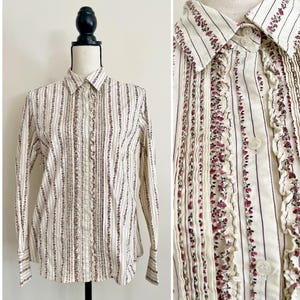 May include: Cream-colored button-down shirt with vertical stripes and floral accents. The long-sleeved shirt features a collared neckline and ruffled detailing down the front. The cuffs have a button closure.