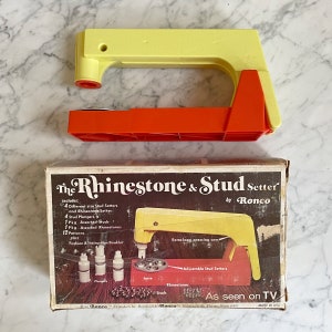 May include: A yellow and red plastic rhinestone and stud setter by Ronco. The box shows the tool with the text "The Rhinestone & Stud Setter by Ronco" and "As seen on TV".
