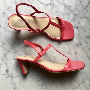 Puede incluir: Un par de sandalias de tacón rosa con diseño de tira en T. Las sandalias tienen puntera cuadrada y tacón de bloque.