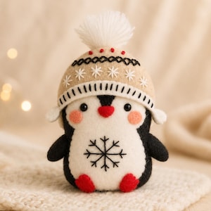 May include: A felt penguin ornament featuring a black and white body, red feet, and a red beak. The penguin wears a tan knit-style hat with a white pom-pom, black and white stitched patterns, and a black snowflake embroidery on its white belly.