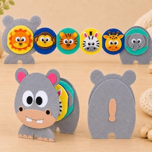 Puede incluir: Un conjunto de juguetes de fieltro con temática animal que incluye un hipopótamo, león, mono, tigre, cebra, jirafa y elefante grises. Las piezas están diseñadas con caras circulares coloridas y cuerpos de fieltro gris, mostrando varios animales.