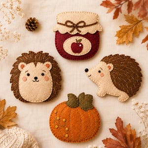 May include: A set of four felt ornaments featuring two hedgehogs, a jar of apple jam with a brown string bow, and an orange pumpkin with green leaves. The hedgehogs are brown and beige with stitched details, while the pumpkin has yellow accents.