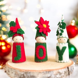 Puede incluir: Tres muñecos de madera cubiertos de fieltro sobre una rodaja de madera circular. Los muñecos visten trajes de fieltro rojo, verde y blanco, cada uno con una letra para deletrear JOY, adornados con acebo, una flor de pascua y muérdago.