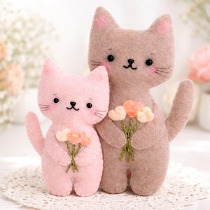 May include: Two felt cat plushies, one smaller light pink and one larger beige, each featuring embroidered black eyes, a small nose, pink cheeks, and whiskers. Both cats are holding a small bouquet of peach-colored flowers with green stems.