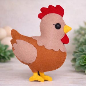 May include: A felt chicken figurine featuring a tan head and wing, a brown body, a bright red comb and wattle, a yellow beak, and yellow feet. The chicken has a single black bead eye and visible stitching along the edges of the felt pieces.