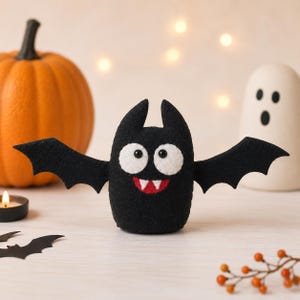 May include: A black felt bat figurine with large white circular eyes, black pupils, and a red mouth featuring two white fangs. The bat has pointed ears and outstretched wings, crafted from soft black felt material with a textured surface.