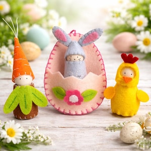 Puede incluir: Tres decoraciones de Pascua de fieltro: una figura con forma de zanahoria con parte superior verde, un conejo gris claro dentro de un huevo de fieltro rosa con una flor y un pollito amarillo con cresta roja, con rasgos faciales pintados.