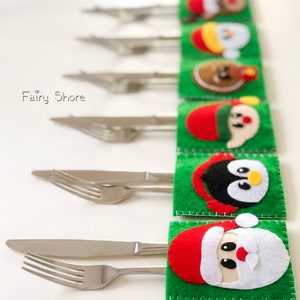 May include: A set of six green felt cutlery holders, each featuring a different holiday character applique including Santa, a penguin, and a gingerbread person. Each holder contains a stainless steel knife and fork. The text Fairy Shore is visible.