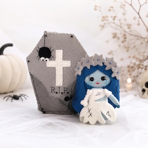 Peut inclure: Un ensemble en feutrine comprenant un cercueil gris avec une croix blanche, l'inscription R.I.P. et une araignée noire, accompagné d'une poupée aux cheveux bleus, une couronne de fleurs grises et une robe blanche avec des détails cousus noirs.