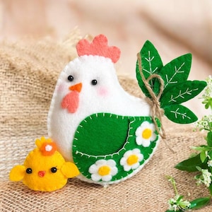 May include: A white felt chicken with a green felt body and a pink felt comb. The chicken has black eyes and a pink beak. The chicken is holding a small yellow felt chick. The chicken is decorated with white felt flowers.