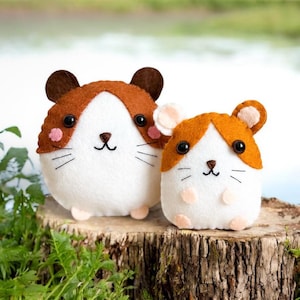 May include: Two felt hamster plushies, one larger than the other, with brown ears and brown accents around their faces. They are both sitting on a tree stump.