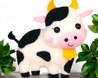 May include: A plush cow toy featuring a white body with black spots, yellow horns, and a yellow bell around its neck. The cow has a pink snout, pink inner ears, pink cheek accents, and black button eyes with a stitched mouth and a black tail.