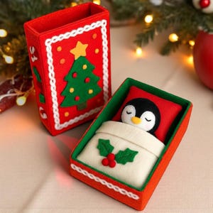 Puede incluir: Una caja navideña de fieltro rojo con un borde blanco y un diseño de árbol de Navidad verde. En el interior, un pingüino de fieltro duerme en una cama en miniatura con una almohada roja y una manta blanca. La caja está decorada con acebo.