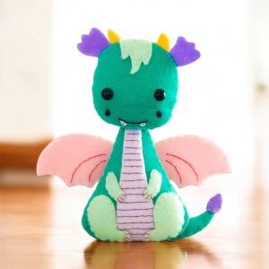 Puede incluir: Un dragón de fieltro verde con alas rosas, cuernos morados y detalles amarillos. El dragón tiene una sonrisa cosida y rayas moradas en la espalda.