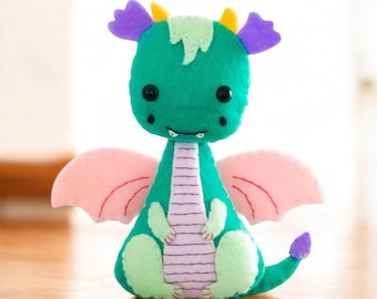 May include: A handcrafted felt dragon plush toy featuring a vibrant teal body, light green belly and feet, and soft pink wings. The creature has purple ear-like accents, yellow horns, and a light green tuft on its head with black bead eyes.
