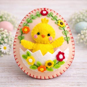 May include: An oval-shaped felt ornament featuring a bright yellow chick emerging from a white cracked eggshell. The design is decorated with embroidered green vines, small red, orange, and yellow felt flowers, and finished with a red blanket stitch border.