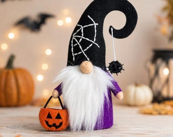 Puede incluir: Una figura decorativa de gnomo con un sombrero alto y curvado de fieltro negro bordado con un patrón de telaraña blanca. El gnomo tiene una larga barba de piel sintética blanca, un cuerpo morado y sostiene un pequeño cubo de calabaza.