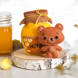 May include: A brown felt teddy bear standing next to a yellow felt honey jar with a brown felt lid and a brown twine bow. The honey jar has a honeycomb design and the word "HONEY" written on it.