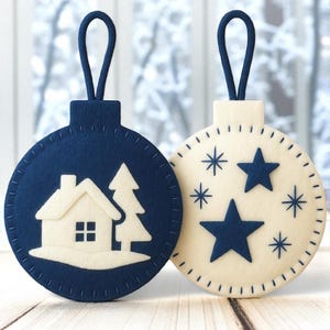 May include: Two felt Christmas ornaments. One is navy blue with a white house and tree design. The other is off-white with navy blue stars and snowflake designs. Both have a navy blue loop for hanging.