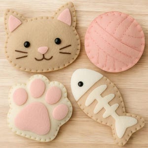 Op de afbeelding: Een verzameling vilten kattenspeeltjes, waaronder een kattenkop, een bol wol, een pootafdruk en een visvormig speeltje. De speeltjes zijn beige en roze, met zwarte en bruine details. De items zijn aan elkaar gestikt.