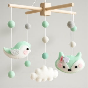 May include: A hanging mobile featuring a light wooden cross frame with white strings suspending felt shapes. The shapes include a mint green bird, a mint green fox head with a white flower, a white cloud, and various mint green and gray felt balls.