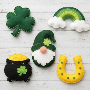 Könnte beinhalten: Eine Sammlung von Filzdekorationen zum St. Patrick's Day. Enthält ein grünes Kleeblatt, einen Regenbogen mit weißen Wolken, einen Wichtel mit rosa Nase, einen Goldtopf und ein gelbes Hufeisen mit grünen Akzenten. Alle Artikel sind mit Kontrastgarn genäht.