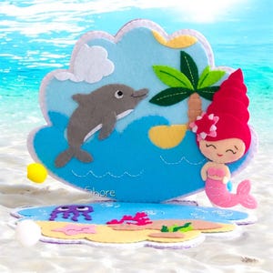 Op de afbeelding: Viltwerk dat een zeemeermin met roze haar en staart, een dolfijn die uit het water springt, een palmboom op een klein eiland en een paarse kwal voorstelt. Het werk is geplaatst tegen een blauwe oceaanachtergrond met witte wolken en het woord "Shore" onderaan.