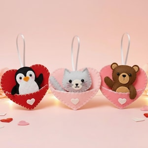 May include: Three felt heart-shaped ornaments with white ribbon loops. The first is a red heart with a penguin, the second is a light pink heart with a grey cat, and the third is a pink heart with a brown bear. Each heart features a small white heart.