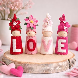 Puede incluir: Cuatro figuras decorativas de madera que deletrean "LOVE", cada una con un diseño diferente de sombrero y flores. Las figuras son rojas, rosas y blancas, y están sobre una rodaja de madera. Hay corazones de fieltro en primer plano.