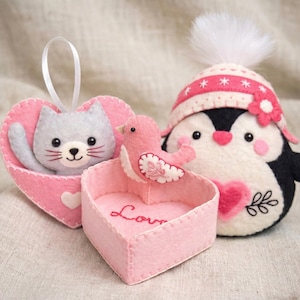 May include: A felt craft set featuring a gray cat inside a pink heart-shaped pouch, a pink bird perched on a heart-shaped box with the word Love embroidered in red, and a penguin wearing a pink hat with a white pom-pom and a pink flower.