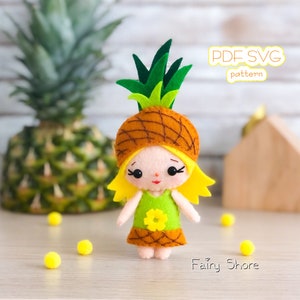 May include: A felt pineapple doll wearing a green dress with a yellow flower. The doll has blonde hair and a smiling face. The doll is standing on a wooden surface with the text "PDF SVG pattern" and "Fairy Shore" visible.