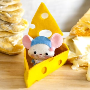 Könnte beinhalten: Eine kleine, weiße Maus mit einem blauen Hut sitzt in einem gelben Filz-Käsekeil. Der Käsekeil hat Löcher, die einem Stück Schweizer Käse ähneln.