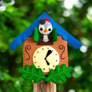 Op de afbeelding: Een vilten koekoeksklok met een blauw dak, bruine wijzerplaat en groene bladeren. De klok heeft een grijze en zwarte vogel met een rode hoed. De wijzers staan ongeveer op 15:00 uur.