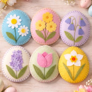 Op de afbeelding: Zes vilten paasei-decoraties in pastelkleuren. Elk ei heeft een ander bloemdesign: madeliefjes, gele bloemen, blauwe klokjes, een hyacint, een tulp en een narcis. De eieren zijn rondom de randen gestikt.