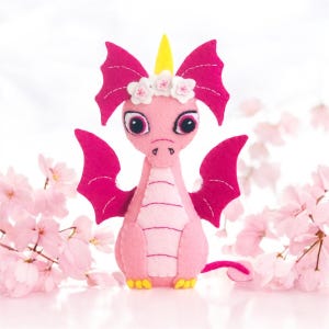 May include: Pink felt dragon with a yellow horn, white wings, and a flower crown. The dragon has large black eyes and a pink body with white stitching.