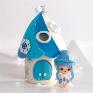 May include: A felt doll of a fairy with blue hair and a white dress stands in front of a white felt house with a blue roof and a blue door. The house has white snowflakes on it.