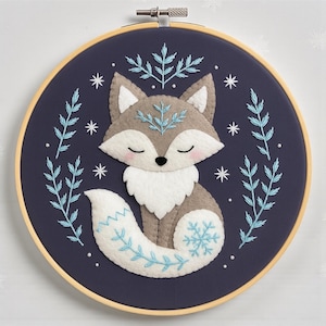 May include: A circular wooden embroidery hoop containing a dark navy fabric background. Centered is a felt fox applique in shades of grey and white with closed eyes and pink cheeks, surrounded by embroidered light blue sprigs and white stars.
