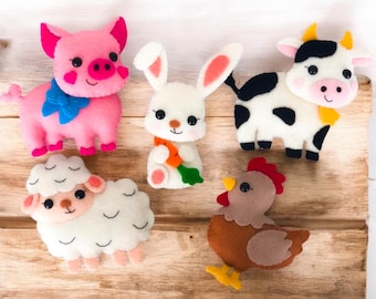 May include: A collection of five felt animal ornaments including a pink pig with a blue bow, a white rabbit holding a carrot, a black and white cow, a white sheep with textured wool, and a brown chicken with a red comb and yellow feet.