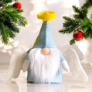 May include: A light blue felt gnome figurine featuring a tall pointed hat topped with a yellow star halo, a long white faux fur beard, and two white felt wings extending from the sides. The gnome has a small peach-colored nose.