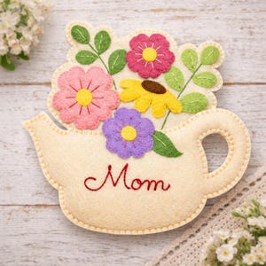 Può includere: Una decorazione in feltro color crema a forma di teiera con la parola Mom ricamata in corsivo rosso. La teiera è decorata con fiori in feltro sovrapposti rosa, gialli e viola, arricchiti da foglie verdi e dettagli cuciti.