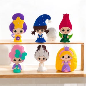 May include: Six felt dolls with different hair colors, outfits, and headwear. The dolls are arranged on a wooden shelf. The dolls are all wearing dresses and have large eyes.