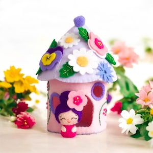 Puede incluir: Una casa de hadas de fieltro con un techo morado y paredes rosas, decorada con flores y hojas. Una pequeña hada de fieltro con cabello morado y un vestido rosa asoma por la ventana.
