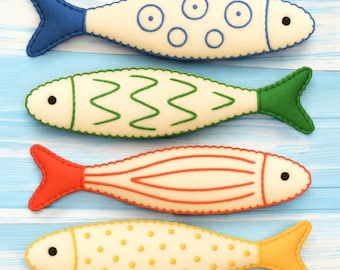 May include: Four felt fish ornaments arranged horizontally. The top fish is cream with blue circles and a blue tail. The second is cream with green zigzag lines and a green tail. The third is cream with orange stripes and an orange tail. The bottom fish is cream with yellow dots and a yellow tail.